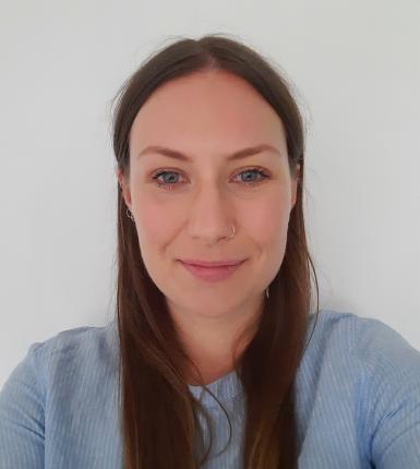 A woman with long brown hair facing the camera, wearing a pale blue long sleeved top.