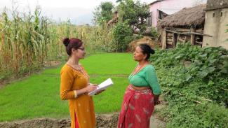 Two women stood facing one another, one is holding a pen and paper. The background to the left is a corn field, and to the right is a house. 