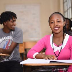 A woman sitting at a desk with a notebook open, ready to study. A man in the background looking at his friend, smiling